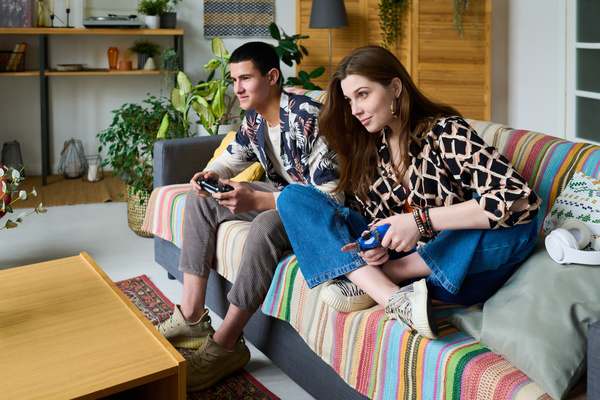 Young friends in bright clothes with prints are sitting on a sofa with a striped bedspread in a bright room with plants and holding joysticks in their hands playing a video game console
